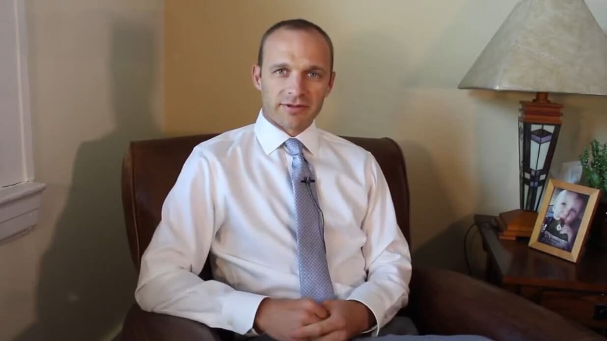 Lakota Denton in his Asheville offices sitting on a brown chair with a yellow wall behind him