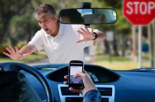 Background is man in white t-shirt about to be hit by vehicle in foreground in shot is steering wheel and hand with a cell phone in the palm